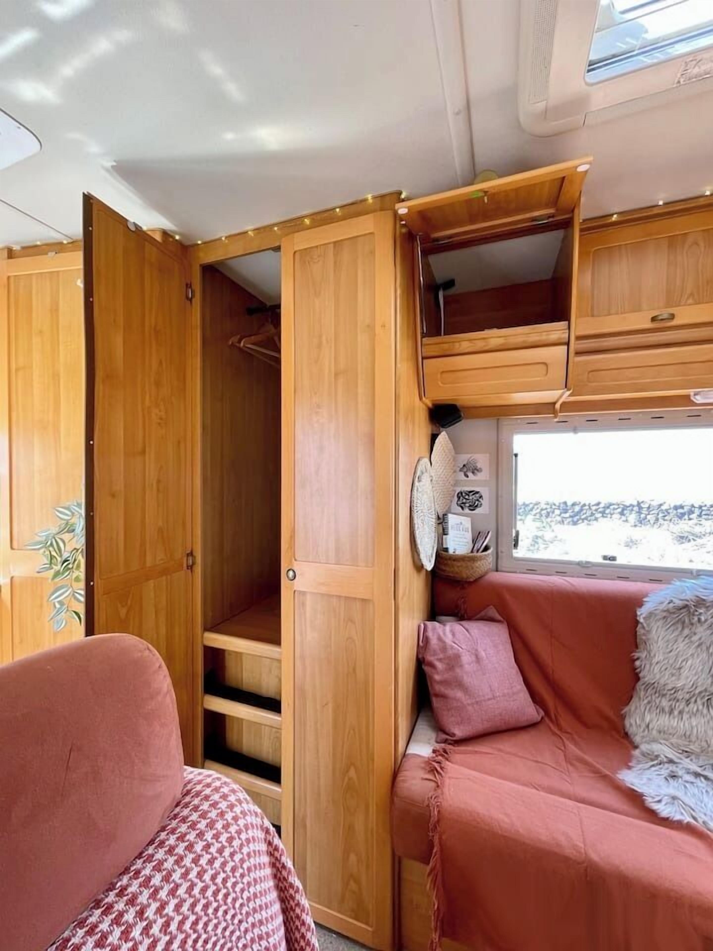 A compact RV interior with a wooden cabinet, a cushioned seating area, and a window providing a view of the outside.