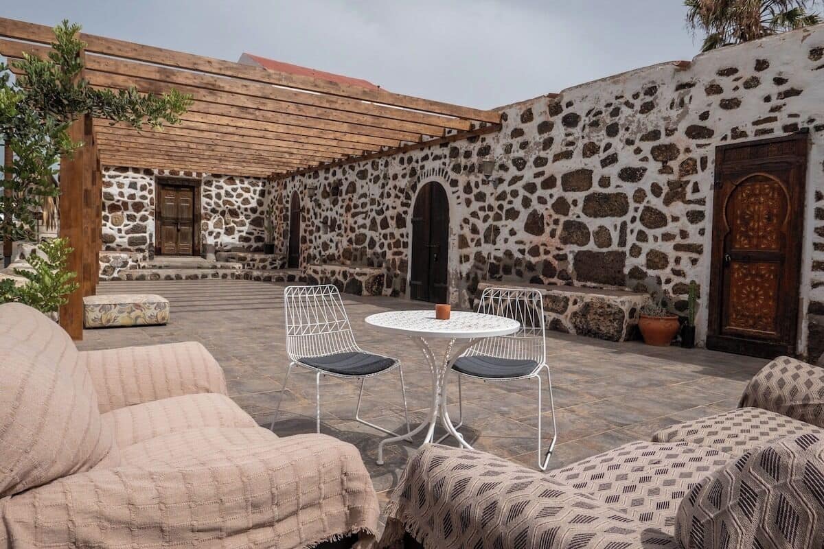 A stone-walled courtyard with a wooden pergola, outdoor seating, and a round table.