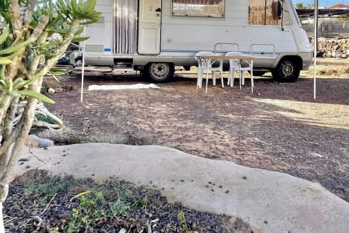 A stationary RV with a covered dining area and a small window, parked in a dry, gravelly area with sparse vegetation.