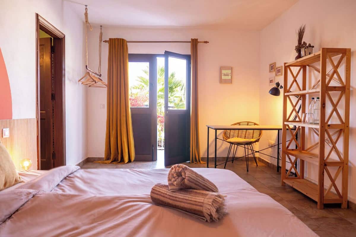 A hotel room with a bed, a desk, a chair, a wardrobe, and a door leading to an outdoor area.