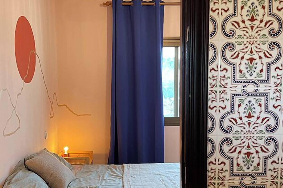 A bedroom with a bed, a bedside table, a lamp, and a window with a curtain.