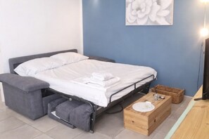 A bed with a grey upholstered bench, a wooden bedside table with a plate, and a wall-mounted artwork.