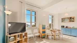 A modern living room with a flat-screen TV, white chairs, a wooden cabinet, and a dining table.