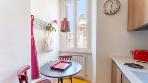 Une petite cuisine avec une table ronde, des chaises, un luminaire suspendu, une horloge et une fenêtre donnant sur des immeubles.