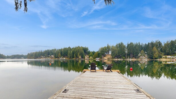 A wooden dock extending into calm water with two chairs facing the water, surrounded by dense forest and houses in the background. - House right on the lake w/ hot tub, kayak, SUP & games - great location (Shelton)