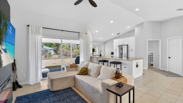 Una sala moderna con un sofá modular, una mesa de centro y un televisor. Se observa también una cocina con armarios blancos y una mesa de comedor.