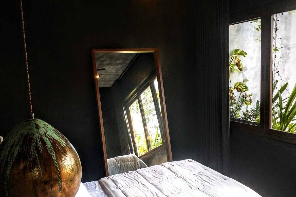A bedroom with a large bed, a hanging light fixture, a mirror, and a window with plants.
