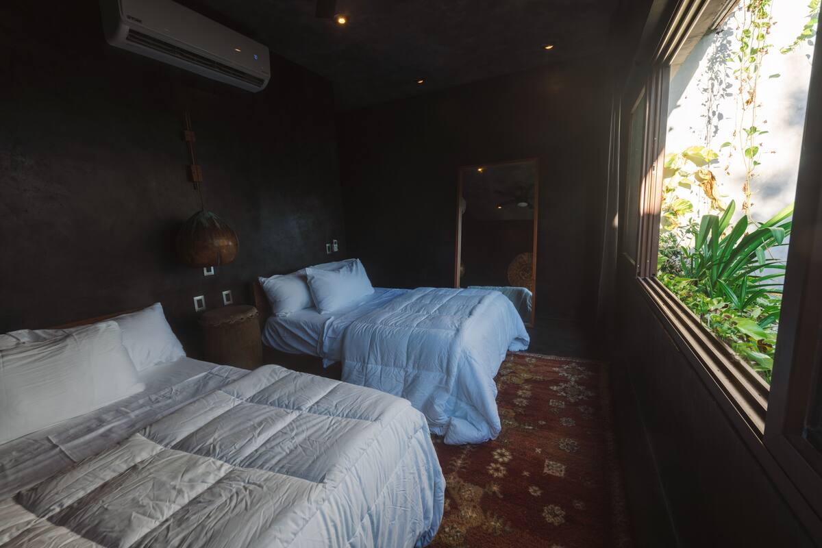 A hotel room with two beds, a red patterned carpet, a large window with plants, and an air conditioning unit.