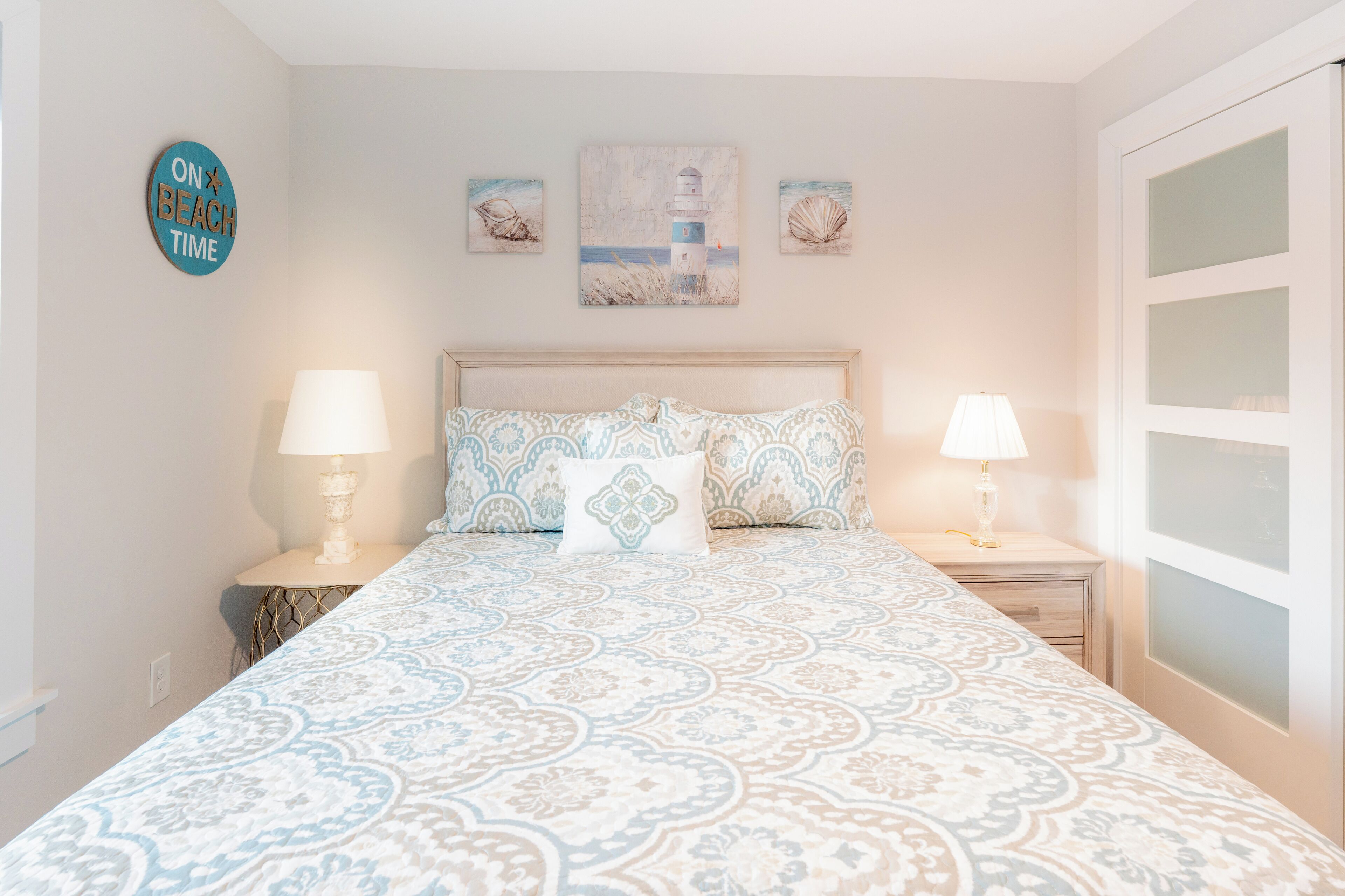 A bedroom with a patterned bedspread, two bedside lamps, a nightstand, and a wall-mounted sign.