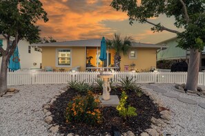 A residential house with a white picket fence, a garden with a birdbath, and a sunset sky.