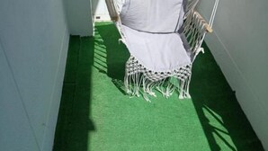 A hammock chair with a white cushion and macrame fringe, set against a green artificial turf floor, against a white wall. - Macarena AL (Almada)