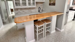 Una cocina con armarios blancos, encimera de madera y un lavabo. Hay dos taburetes con asientos de mimbre.