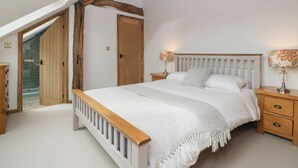 Une chambre avec un plafond en pente, des poutres en bois, un lit avec une literie blanche, une table de chevet avec une lampe et une porte donnant sur une autre pièce.