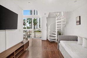 A modern interior with a flat-screen TV, a sofa, a staircase, and a view of palm trees and buildings.