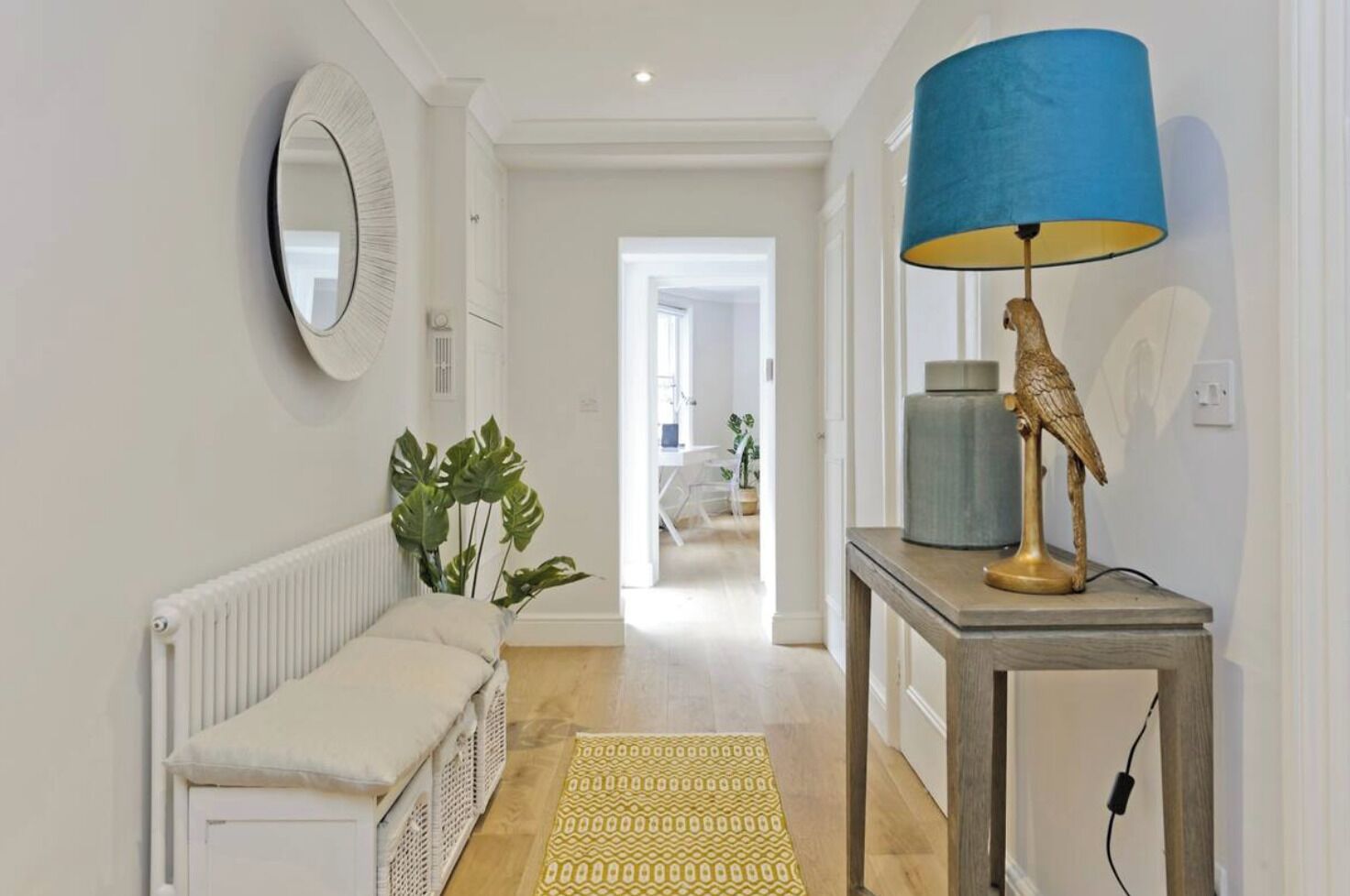 A hallway with a white radiator, a beige bench, a green plant, a yellow patterned rug, and a table with a blue lamp and a bird sculpture.