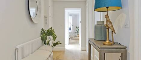 A hallway with a white radiator, a beige bench, a green plant, a yellow patterned rug, and a table with a blue lamp and a bird sculpture.