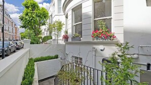 A white townhouse with a curved facade, a small balcony with potted plants, and a narrow front garden. - Stunning garden apartment in heart of Kensington (London)