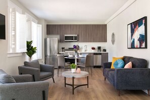 A modern living room with grey sofas, a round coffee table, and a dining area with a white table and chairs. A kitchen with a microwave and stainless steel appliances is visible in the background. - The Penn Hotel - Central 2BR Hotel Suite Stay in Historic Gaslamp (San Diego)