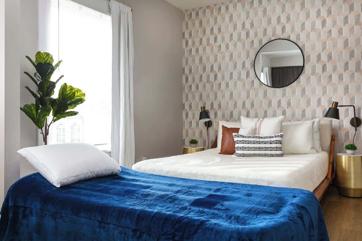 Une chambre à coucher avec un lit, une grande fenêtre, une plante en pot et un mur orné d’un motif géométrique.