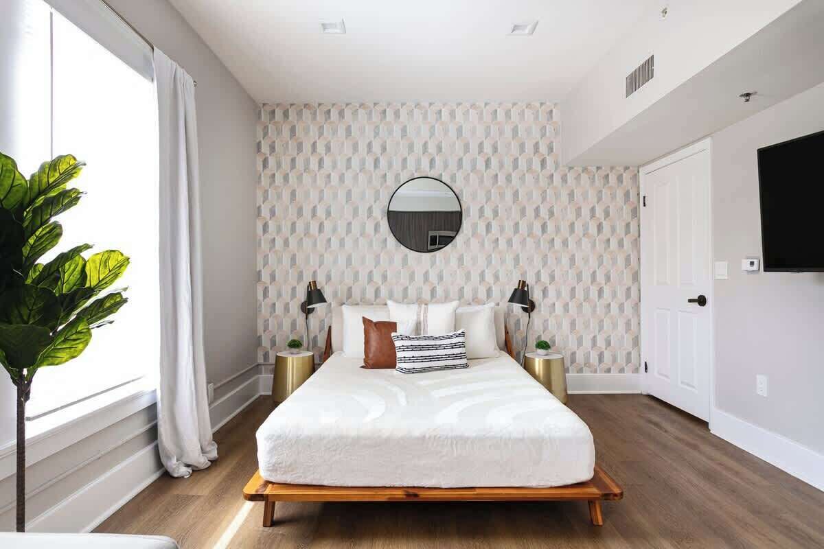 Une chambre à coucher moderne avec un lit en bois, une literie blanche, un miroir rond et un mur orné d’un motif géométrique.