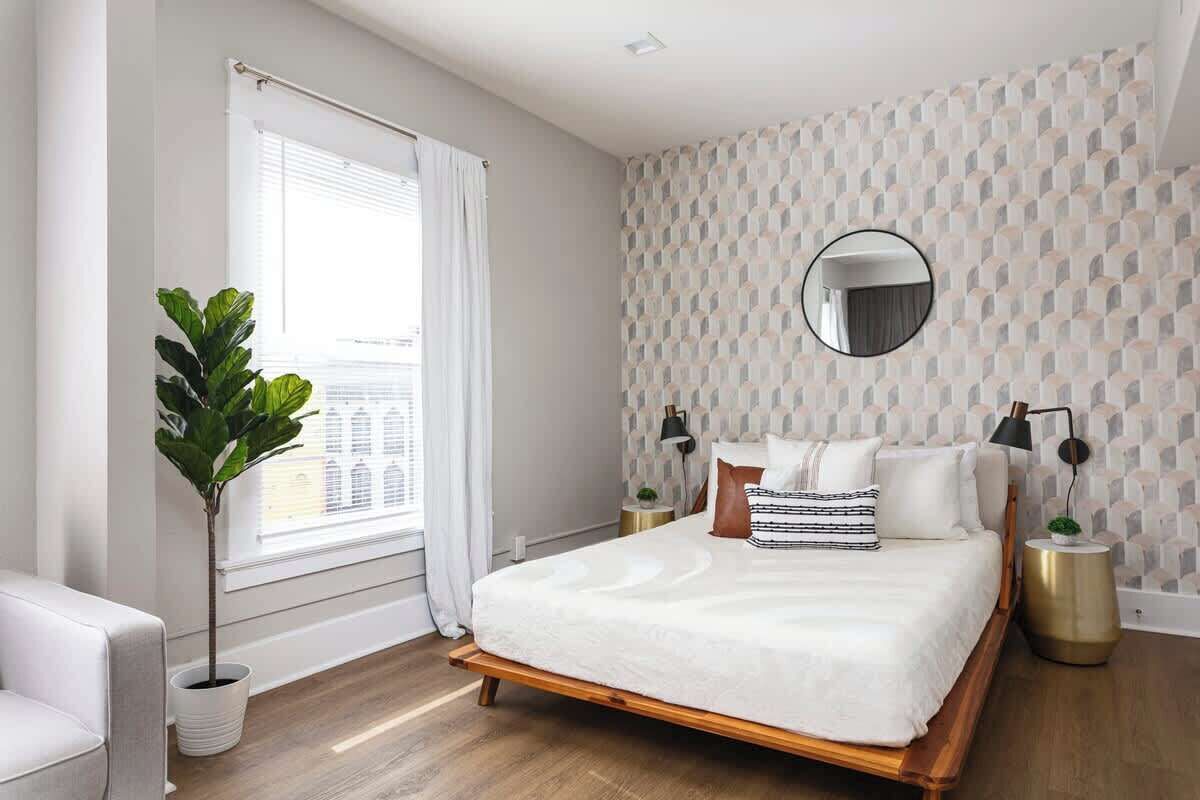 A bedroom with a wooden bed, white bedding, a round mirror, two bedside tables with lamps, a potted plant, and a window with curtains.