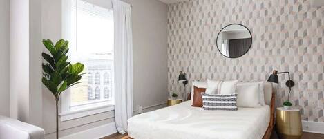 A bedroom with a wooden bed, white bedding, a round mirror, two bedside tables with lamps, a potted plant, and a window with curtains.