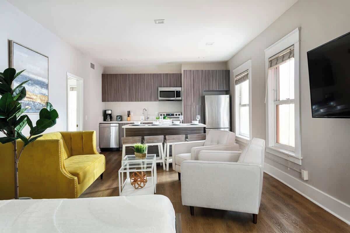 A modern living room with a yellow sofa, white armchairs, and a coffee table. A kitchen with a microwave and refrigerator is visible in the background.