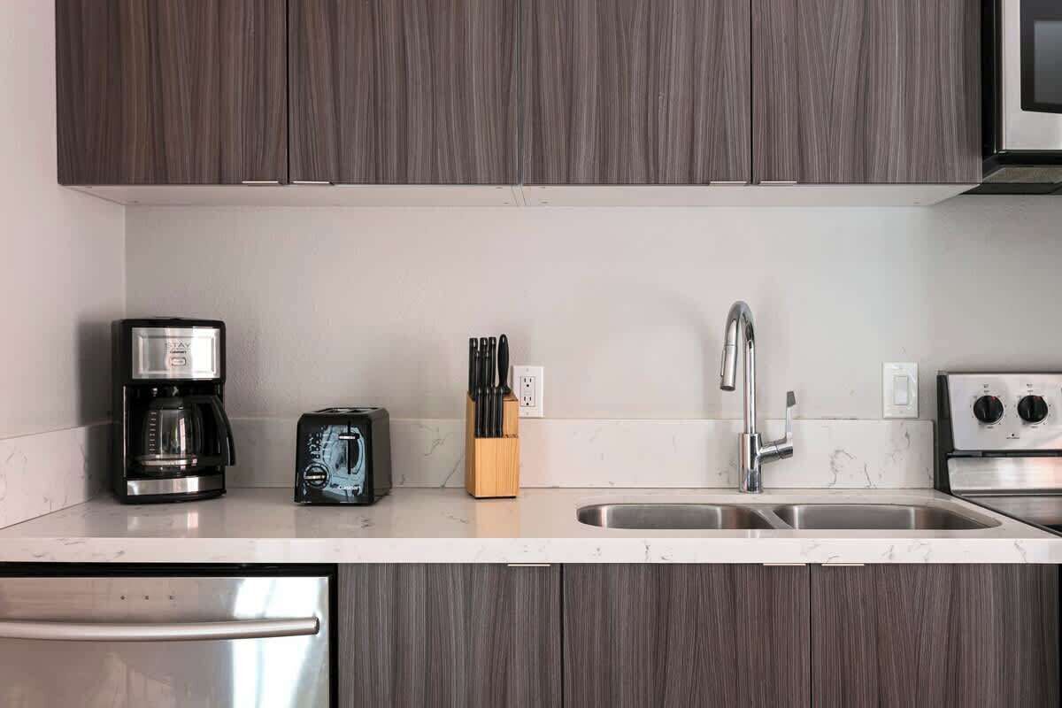A modern kitchen with dark wood cabinets, a stainless steel refrigerator, a coffee maker, a toaster, a knife block, a sink with a faucet, and an oven.