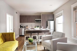 A modern kitchen with a central island, white chairs, a yellow sofa, and a large window.