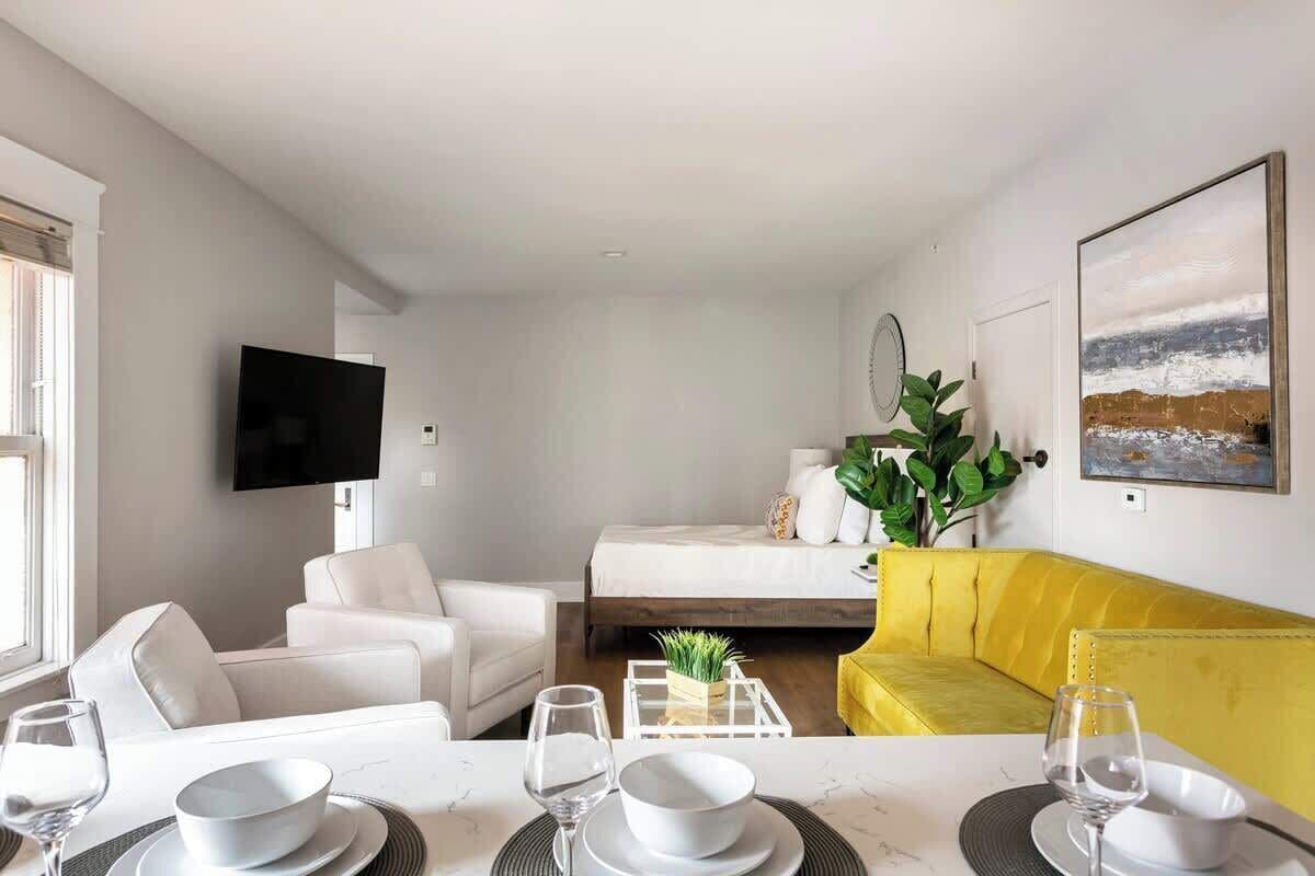Uma sala de estar moderna com um sofá, uma mesa de jantar posta com pratos e copos, uma cama com detalhes amarelos e uma televisão fixada na parede.