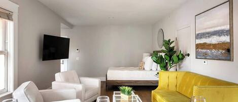 A modern living room with a sofa, a dining table set with plates and glasses, a bed with a yellow accent, and a TV mounted on the wall.