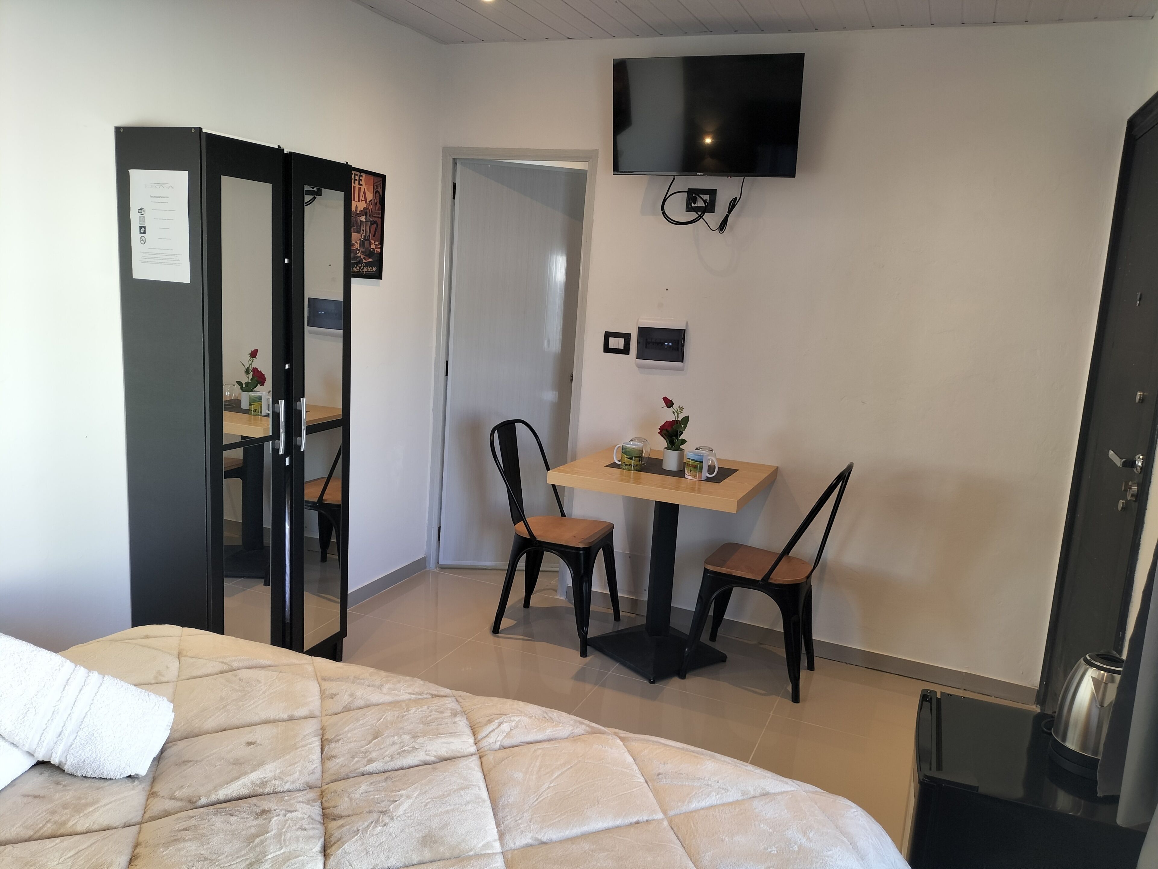 Hotellihuone, jossa on sänky, seinälle asennettu televisio, pieni pöytä tuoleineen ja kahvinkeitin.