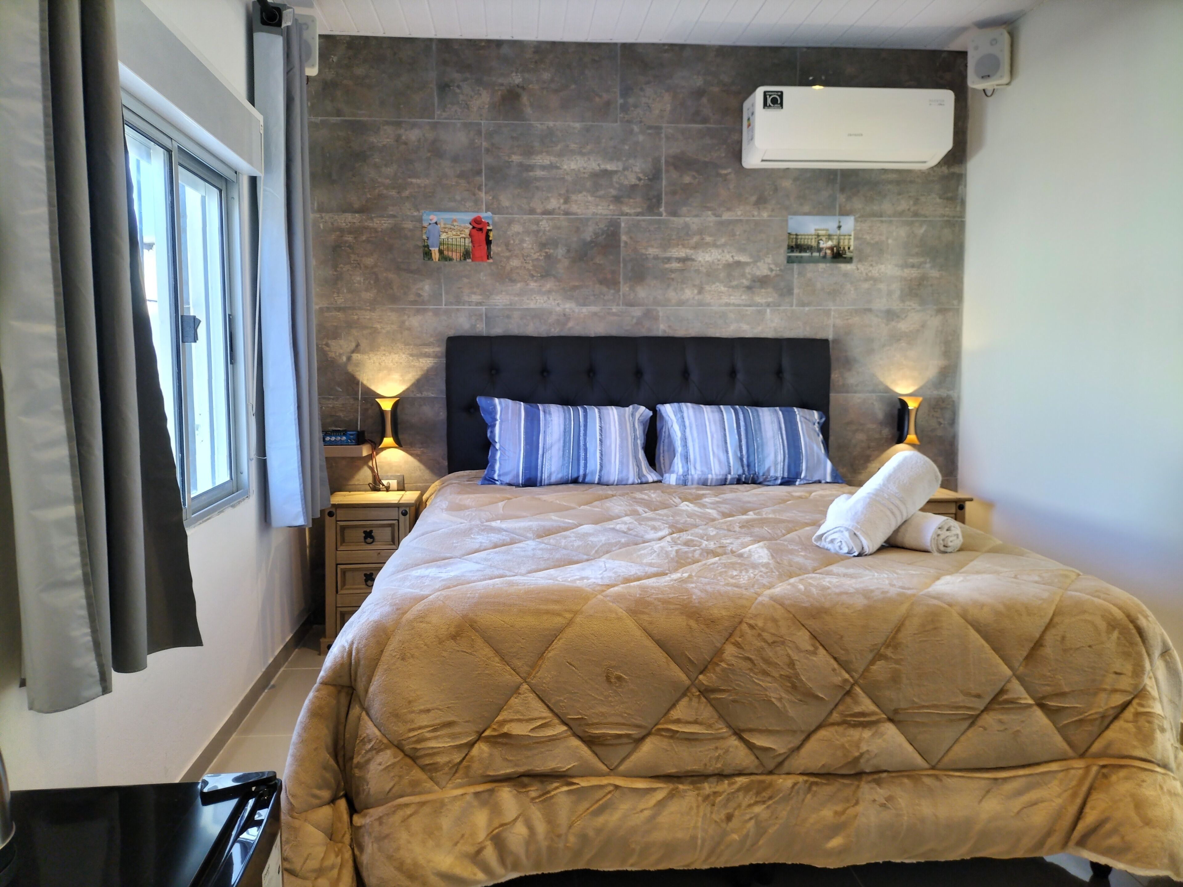 A hotel room with a large bed, two bedside lamps, a window with curtains, an air conditioning unit, and a wall-mounted picture.