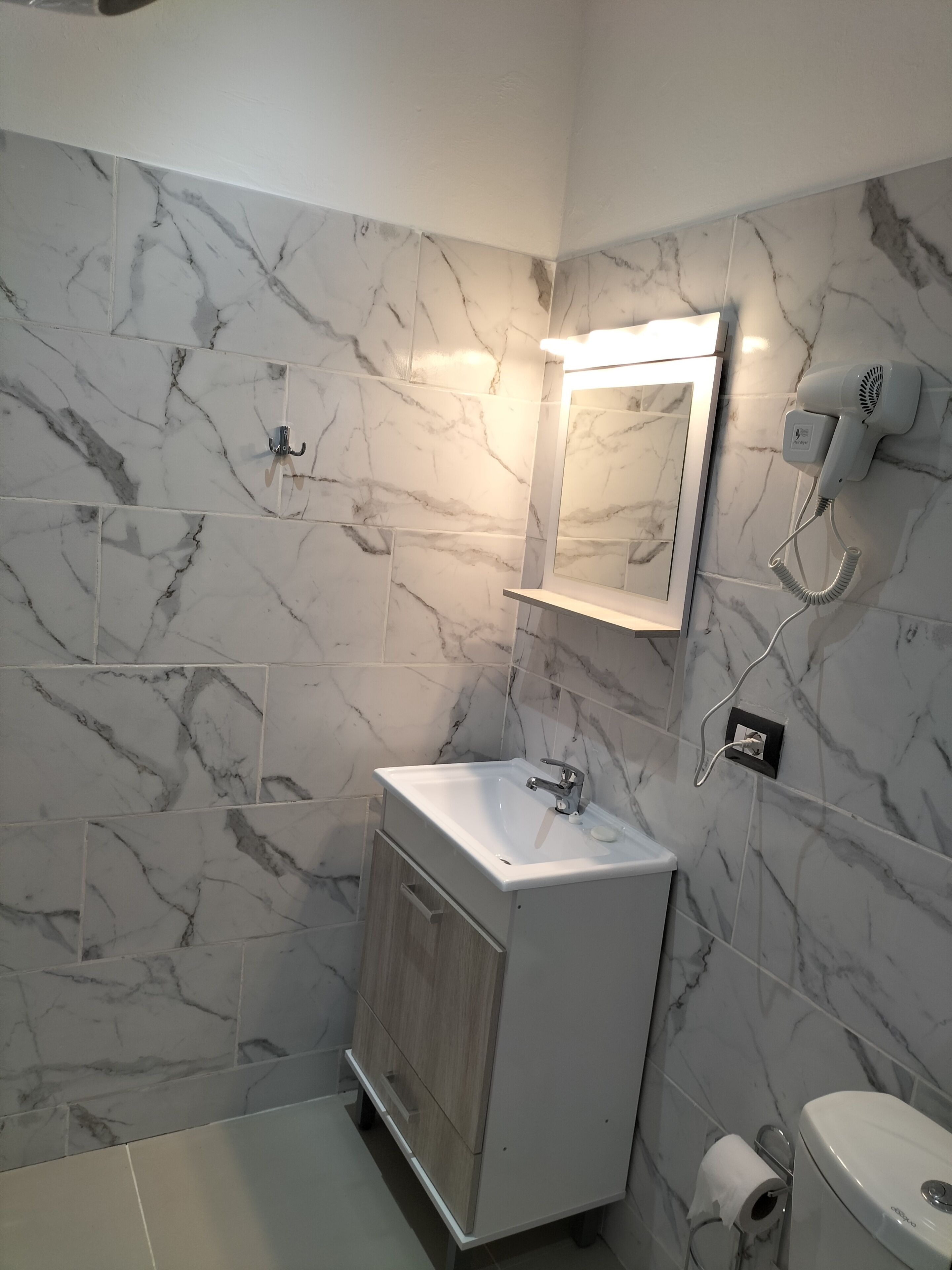 A bathroom with a marble wall, a sink, a mirror, a hairdryer, and a toilet.