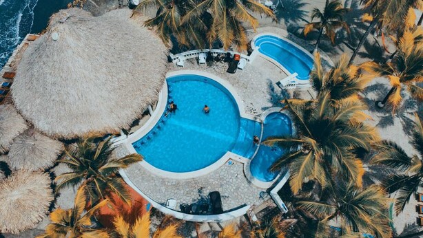 Oversigtsbillede af et resort med en central swimmingpool omgivet af palmer og stråtagparasoller.