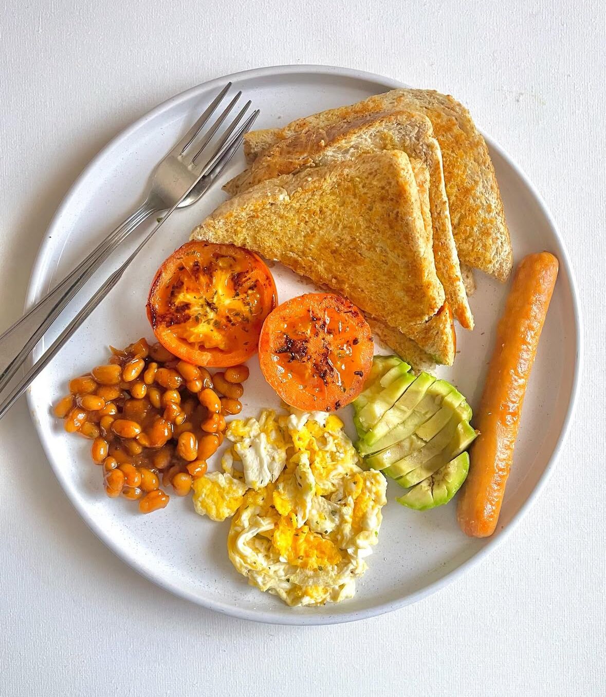 Ein Teller mit Toast, Rührei, Bohnen, Tomaten, Avocado und einer Wurst.