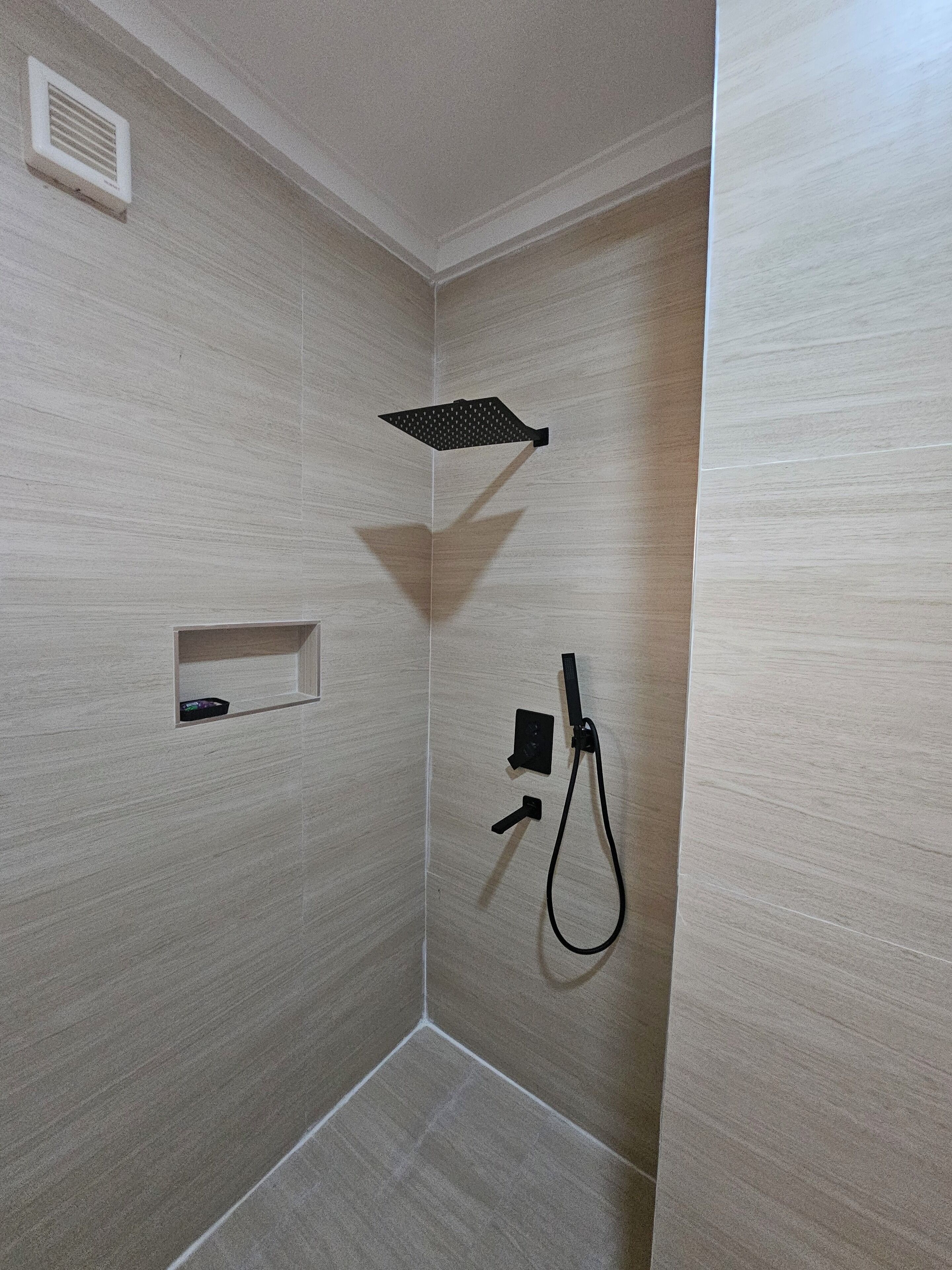 A small, enclosed shower area with a handheld showerhead, a control panel, and a storage shelf.
