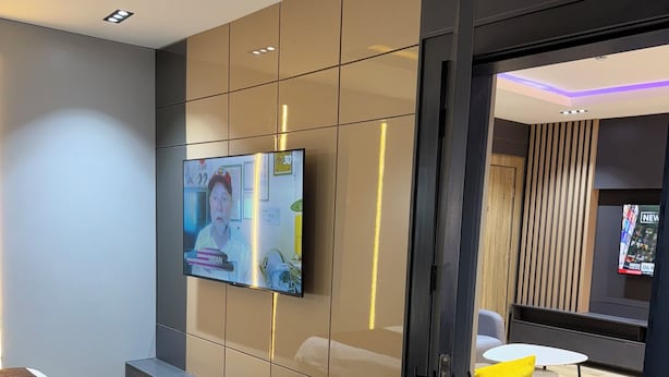 A modern hotel room with a large flat-screen TV mounted on a wall, a wooden bench, a small round table with a yellow pillow, and a bed with a dark cover. - Sapphire Suite (Abuja)