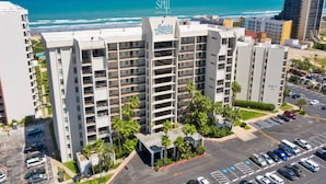 A high-rise hotel with multiple balconies, a parking lot, and a view of the ocean.