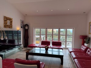 A spacious living room with a large glass coffee table, red leather sofas, a TV, and a view through large windows.