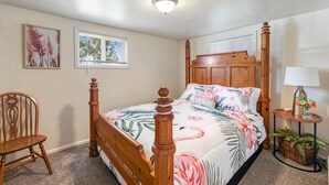 Un dormitorio con cama de madera, una silla, una mesita con lámpara y dos ventanas.