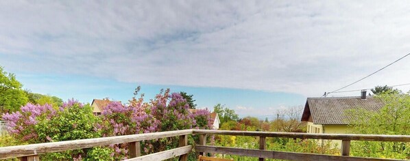 A wooden bridge leading to a house with a tiled roof, surrounded by blooming purple flowers.