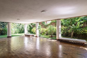 An open-air covered patio with tiled flooring, white columns, and a view of lush greenery.