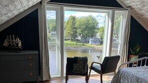 Ein Schlafzimmer mit schrägem Dach, einem Bett, einem Sessel, einer Kommode und einem Fenster mit Seeblick.