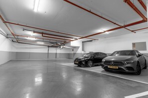A parking garage with two cars, white walls, and visible red pipes on the ceiling. - Luxury 3 rooms Apt• Balcony• Parking | HaKerem A23 (Tel Aviv)