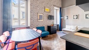 Een kamer met een bakstenen muur, een eettafel met stoelen, een bank, een televisie en kunstwerken aan de muur.