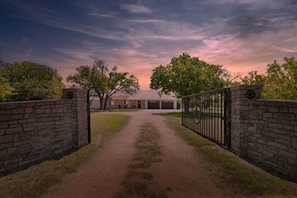 A gated driveway leading to a building with a garage, surrounded by trees and a clear sky. - Rivers Oaks-The Ranch House Pool & Hot-Tub! (Fredericksburg)