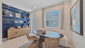 Un coin repas avec une table ronde, des chaises en osier, un mur bleu avec des étagères et une fenêtre avec des stores.