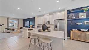 A modern kitchen with a central island, bar stools, a refrigerator, and a dining area with a sofa and coffee table. - New! Waterside Lakewood Ranch, Tennis, Pool, Walk to Restaurants and More! (Sarasota)