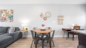 A modern living room with a gray sofa, a wooden dining table with four chairs, a small side table with a lamp, and a wall-mounted macrame piece. - Unit #606 Summary:<br>19 (Clarksville)